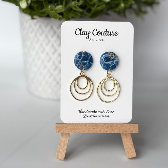 Polymer clay marbled earrings in Midnight blue  color with resin finish and brass charms on a 'Clay Couture' branded card with a plant in the background.