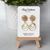 Polymer clay marbled earrings in warm sandstone color with resin finish and brass charms on a 'Clay Couture' branded card with a plant in the background.