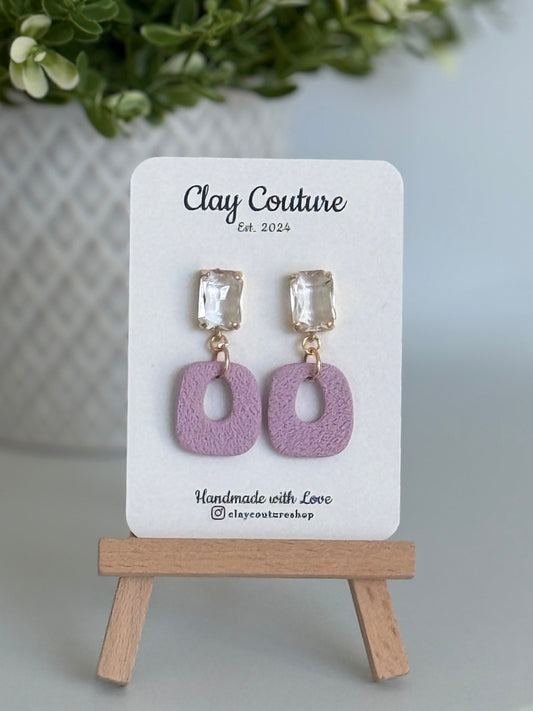 Mauvepolymer clay earrings with a crystal stud on a 'Clay Couture' branded card with a wooden stand.