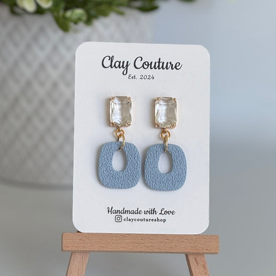 Dusty Blue polymer clay earrings with a crystal stud on a 'Clay Couture' branded card with a wooden stand.