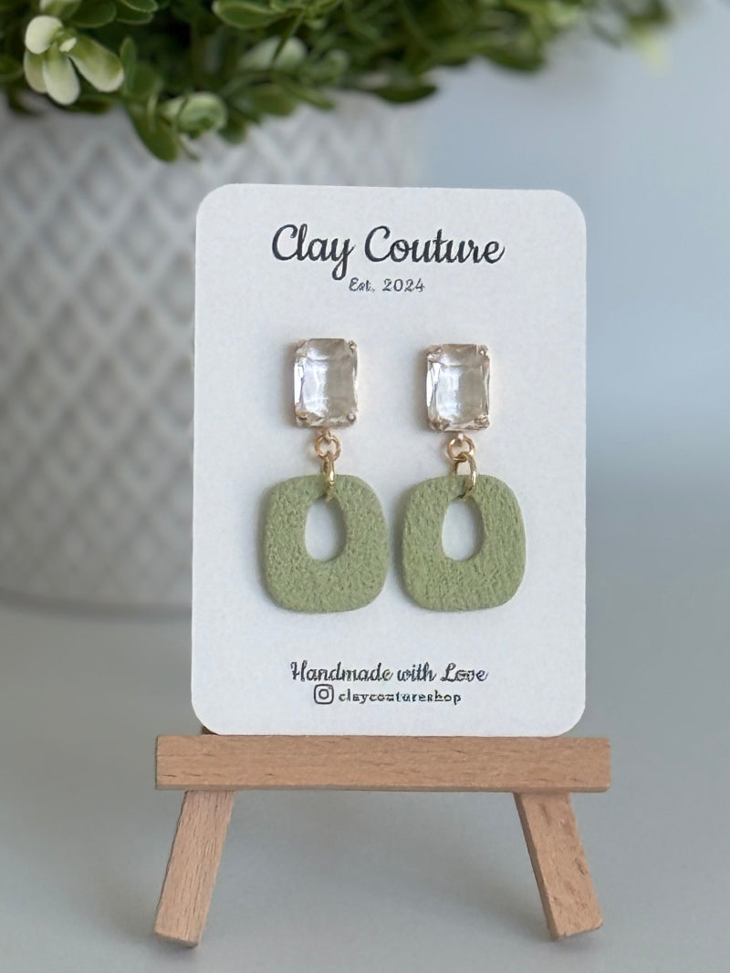 Sage polymer clay earrings with a crystal stud on a 'Clay Couture' branded card with a wooden stand.