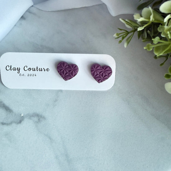 Purple heart-shaped earrings on a white card with 'Clay Couture' branding, placed on a marble surface.