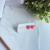 Pink heart-shaped earrings on a white card with 'Clay Couture' branding, placed on a marble surface with greenery.