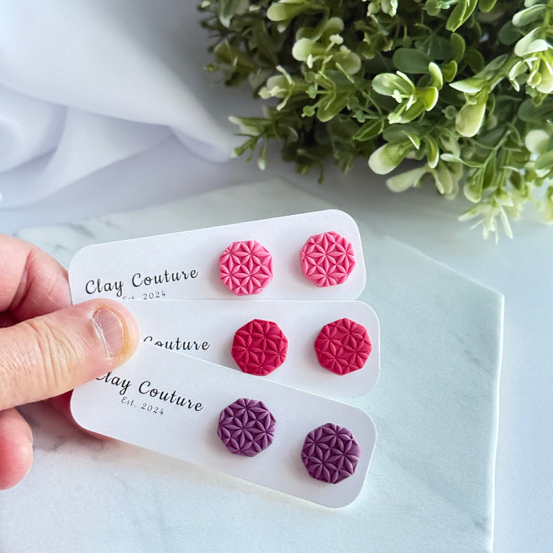 Set of pink and purple floral earrings on a 'Clay Couture' branded card with greenery in the background.