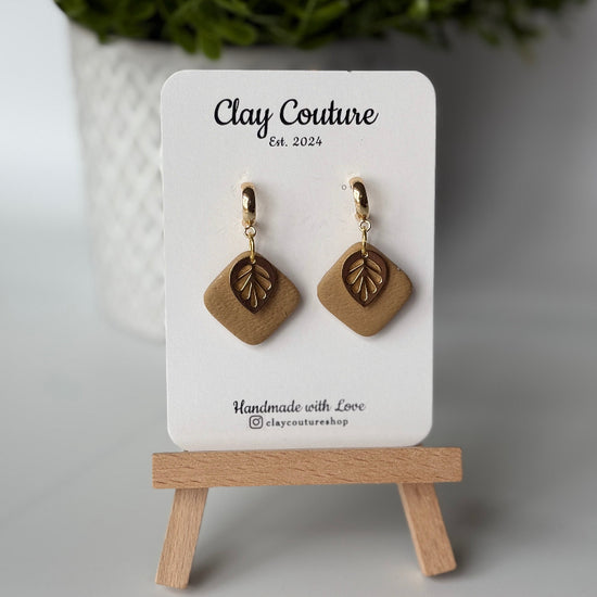 Polymer clay  earrings in warm sandstone color with and gold huggies brass charms on a 'Clay Couture' branded card with a plant in the background.