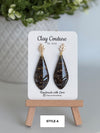 Black teardrop earrings on a 'Clay Couture' branded card with a wooden stand.
