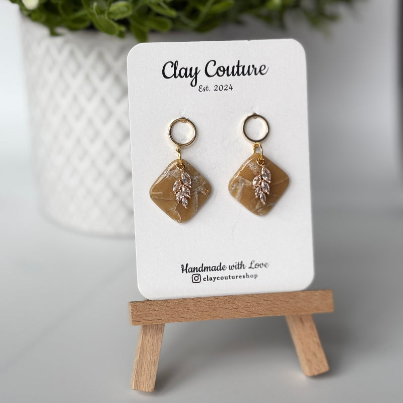 Pair of marbled polymer clay earrings with resin finish  in warm sandstone with crystal charm and gold plated studs on Clay Couture branded card on a wooden stand