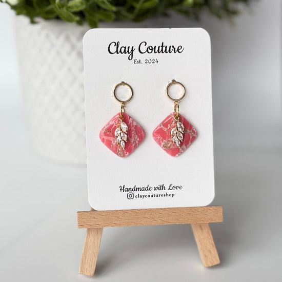 Pair of marbled polymer clay earrings with resin finish  in blush with crystal charm and gold plated studs on Clay Couture branded card on a wooden stand