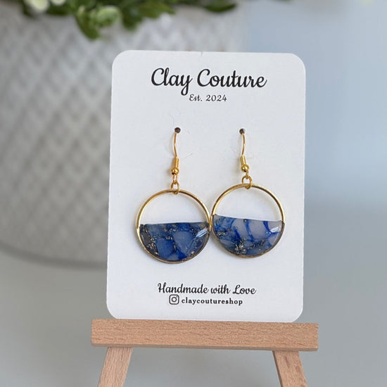 Pair of blue and gold earrings on a 'Clay Couture' branded card with a wooden stand.