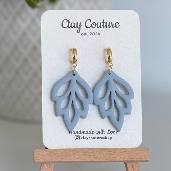 Embossed leaf design  earrings in Duty Blue color with gold plated hoops on a 'Clay Couture' branded card with a plant in the background.