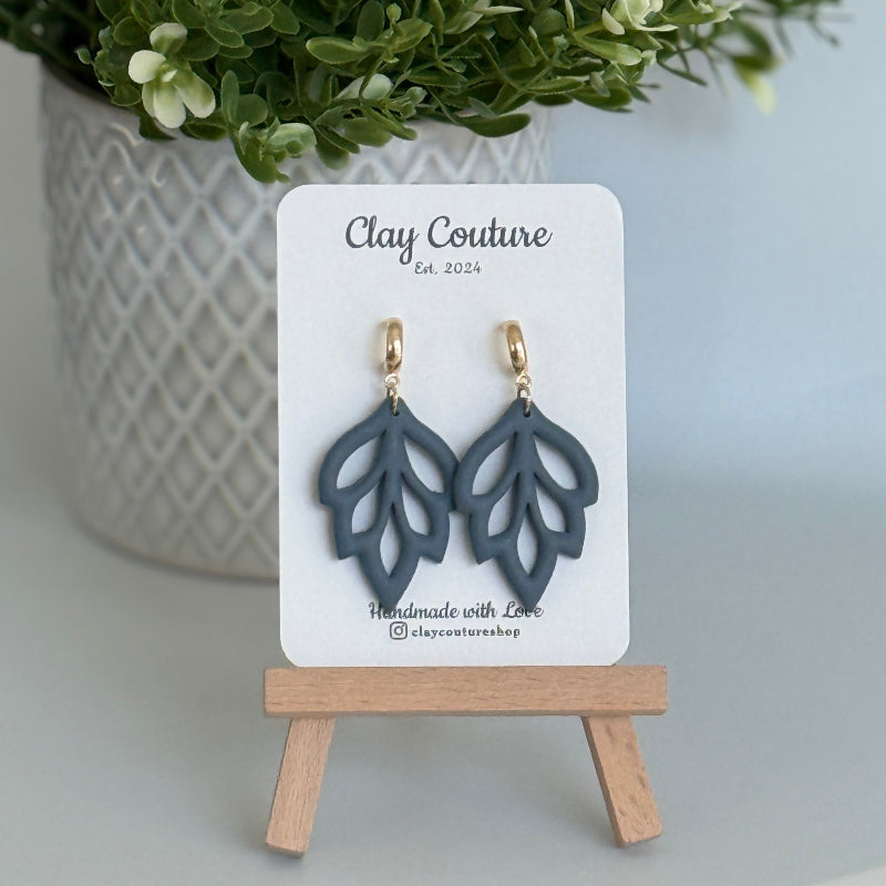 Embossed leaf design  earrings in Slate Blue color with gold plated hoops on a 'Clay Couture' branded card with a plant in the background.