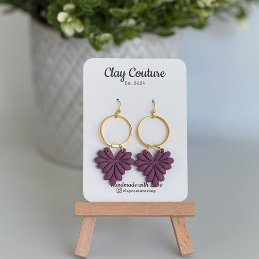 Leaf earrings  in Plum color made out of polymer clay with hypoallergenic attachments on a Clay Couture branded card with a plant in the background.