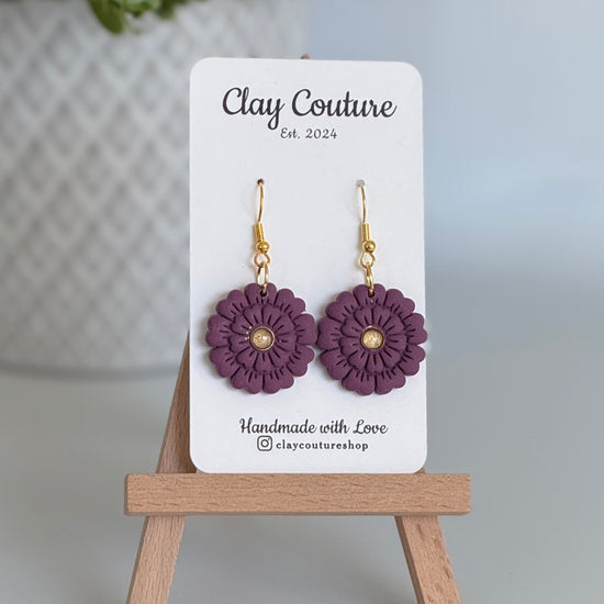 Plum flower earrings on a Clay Couture branded card with a wooden stand.