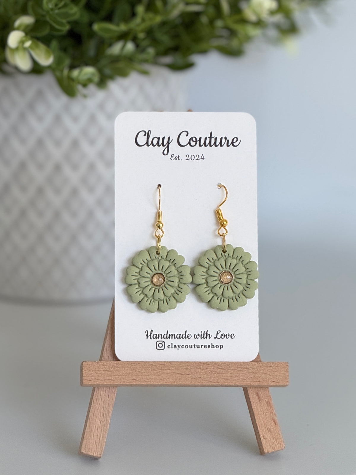 Sage flower earrings on a Clay Couture branded card with a wooden stand.