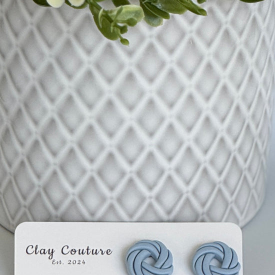 Dusty Blue Polymer Clay knot earrings on a Clay Couture card with a potted plant in the background