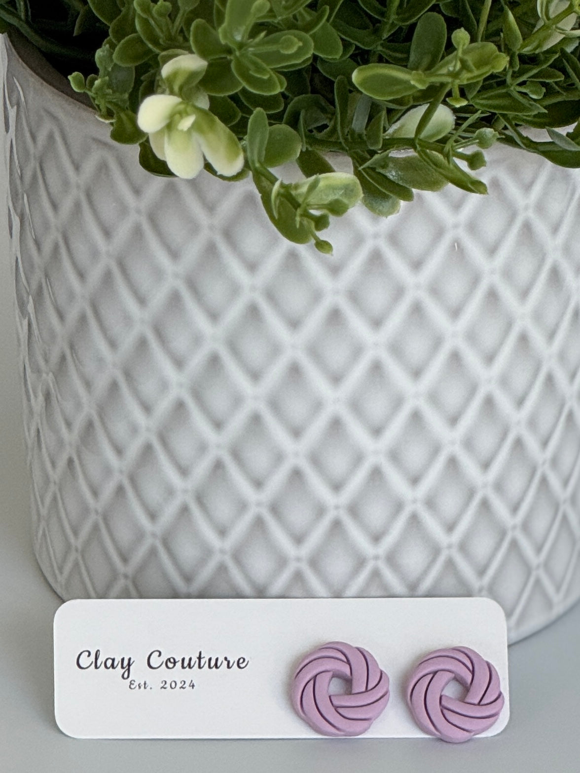 Mauve Polymer Clay knot earrings on a Clay Couture card with a potted plant in the background.