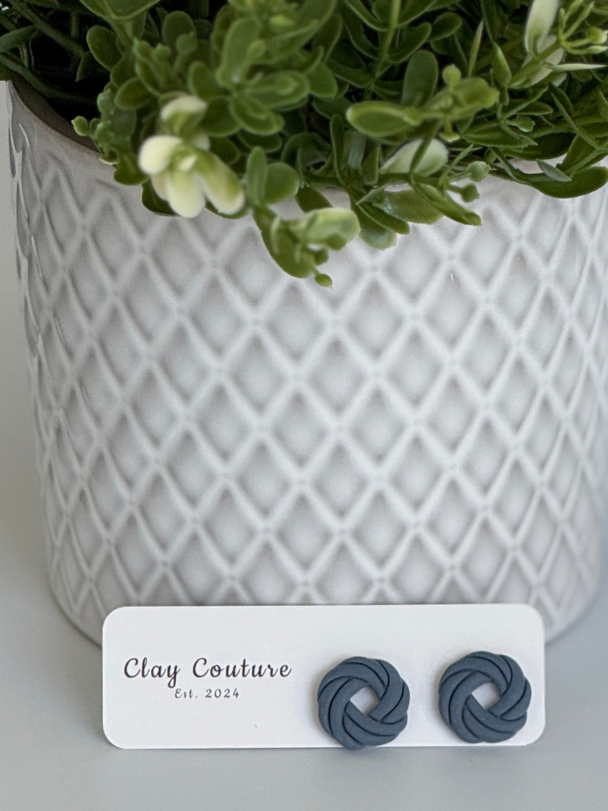 Slate Grey Polymer Clay knot earrings on a Clay Couture card with a potted plant in the backgroud.