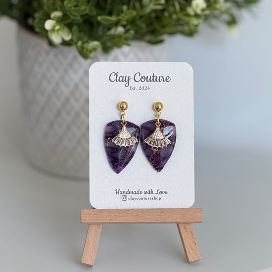 Amethyst purple dagger shaped polymer clay  earrings  with a resin finish and gold accents on a Clay Couture branded card 