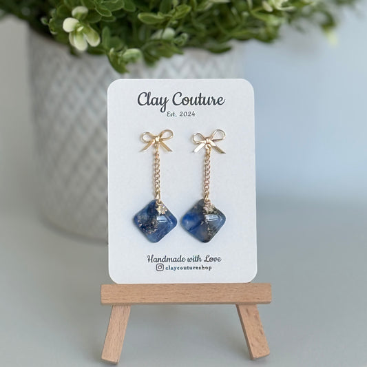 Sapphire Blue earrings  with a gold chain and bow stud on a 'Clay Couture' branded card with a plant in the background
